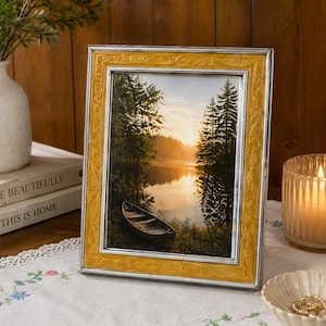 May include: A rectangular picture frame with a metallic silver inner border and a wide, textured mustard yellow outer border. The frame contains a photograph of a canoe on a calm lake surrounded by pine trees at sunrise.