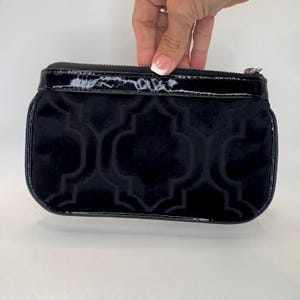 May include: A black velvet clutch bag with a geometric pattern and a glossy black patent leather trim. The bag has a zip closure and a rectangular shape. The bag is held by a hand.