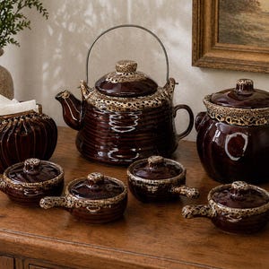 May include: A collection of brown ceramic kitchenware with a speckled beige trim. The set includes a teapot, a covered casserole dish, several small lidded bowls with handles, and a napkin holder. The items are arranged on a wooden surface.