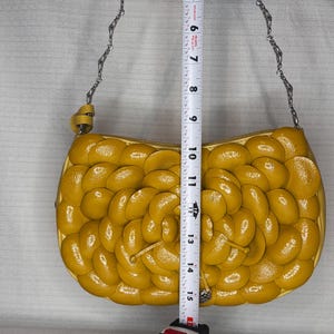Może przedstawiać: Musztardowa torebka z kwiatowym wzorem. Torebka ma srebrny łańcuszek i mierzy około 33 cm długości. Torebka jest wykonana z błyszczącego materiału i ma zapięcie na zamek błyskawiczny.
