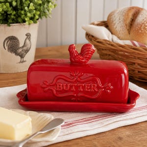 May include: A bright red ceramic butter dish with a rectangular base and a matching lid. The lid features a sculpted rooster handle on top and the word BUTTER embossed in the center within a decorative frame. The dish has a glossy finish.