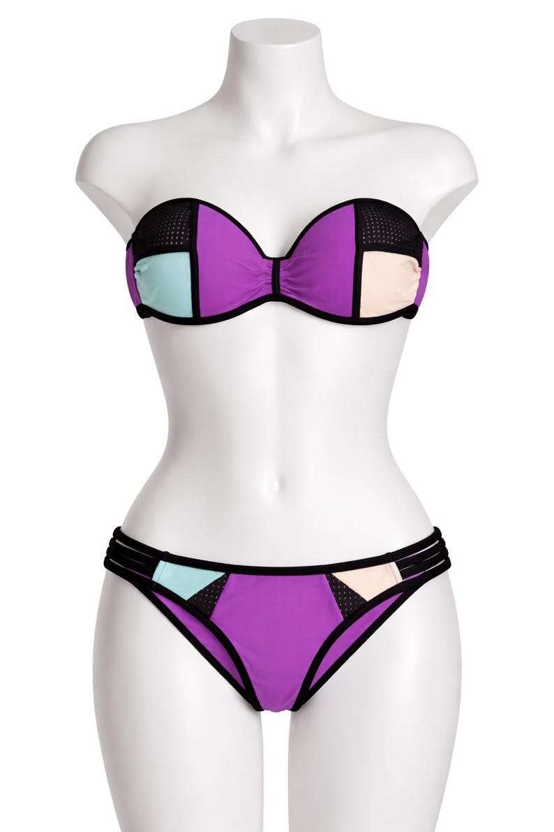 May include: A two-piece bikini set on a mannequin. The top is a strapless design with a purple base, black mesh accents, and colour block details in light blue and peach. The bottoms are purple with matching colour block details.