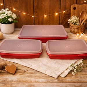May include: A set of three red rectangular food storage containers with light pink lids. The containers are arranged on a wooden surface with a white cloth, flowers, and decorative lights in the background. The lids have a circular design.