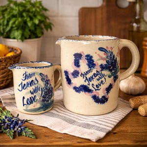May include: A cream-colored ceramic pitcher and mug set. The pitcher and mug are decorated with blue and pink floral designs and the words "From Texas with Love!" The set is on a striped cloth.
