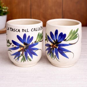 May include: Two white ceramic cups with a blue floral design and green leaves. The cups have the text "LA TASCA DEL CALLEON" in black. The cups are on a white surface.