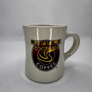 May include: Cream-colored ceramic coffee mug with a curved handle. The mug features the "WAFFLE HOUSE COFFEE" logo in a circular design with red, yellow, and black colors. The mug has a slightly flared shape and a glossy finish.