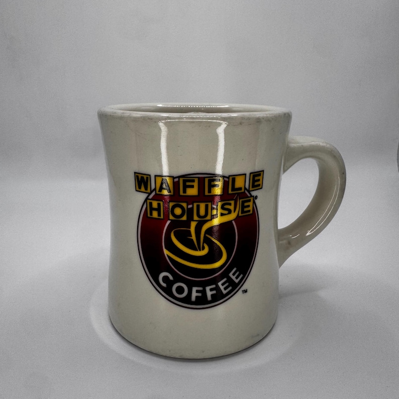 May include: Cream-colored ceramic coffee mug with a curved handle. The mug features the "WAFFLE HOUSE COFFEE" logo in a circular design with red, yellow, and black colors. The mug has a slightly flared shape and a glossy finish.