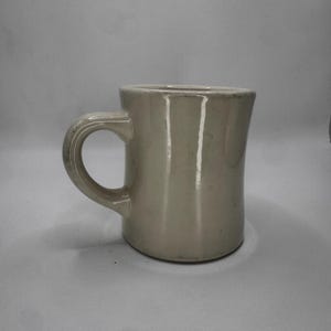 May include: A cream-colored ceramic mug with a rounded handle. The mug has a simple, classic design, suitable for coffee or tea. The mug is made of a sturdy material and is likely dishwasher safe.
