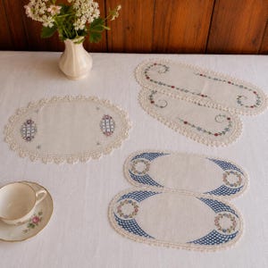 May include: A collection of five oval-shaped linen doilies with intricate embroidery and scalloped lace edges. The pieces feature blue grid patterns and floral motifs. A white ceramic teacup with a floral design sits on a matching saucer.