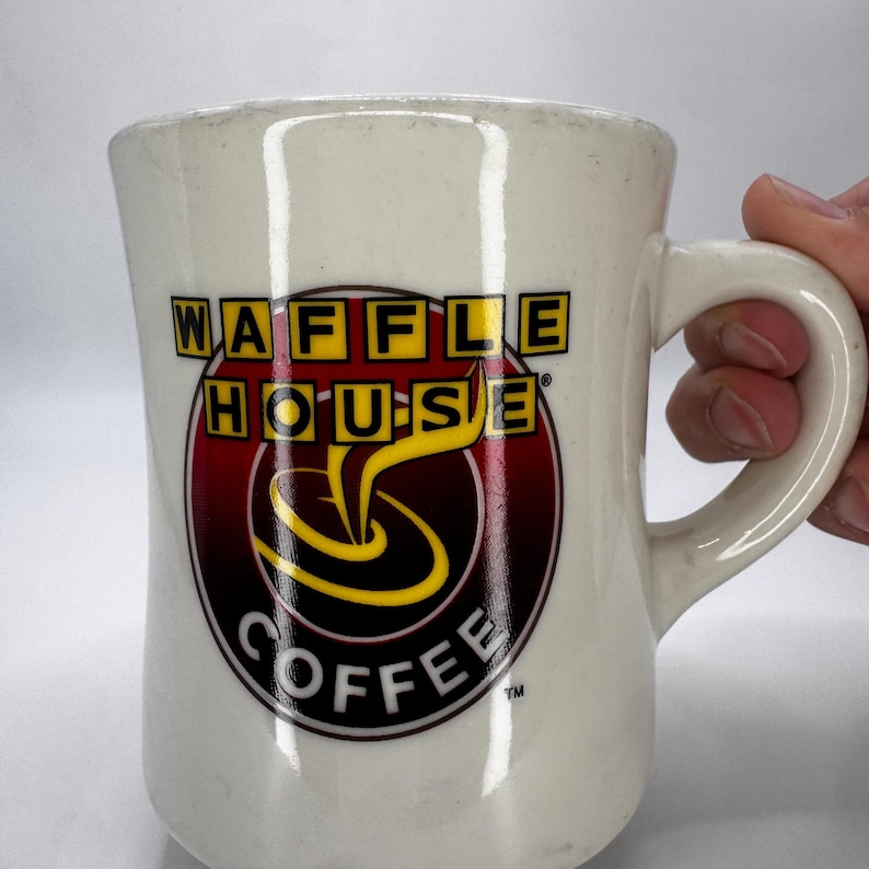 May include: Cream-colored ceramic coffee mug with the "WAFFLE HOUSE COFFEE" logo in yellow, red, and black. The mug has a curved handle and a slightly flared top. The logo features a stylized coffee cup design.