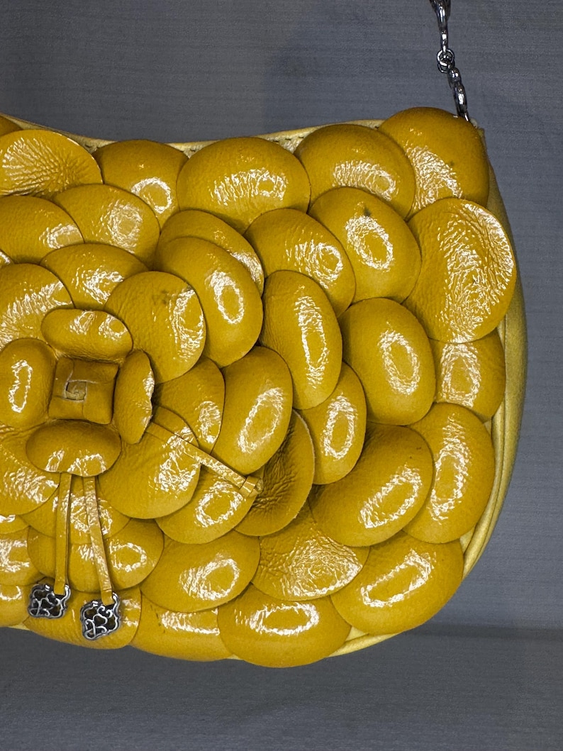 Może przedstawiać: Musztardowa torebka z unikalnym, teksturowanym wzorem. Torebka posiada szereg zachodzących na siebie, owalnych element&oacute;w, tworzących wz&oacute;r przypominający łuski. Srebrny łańcuszek i dekoracyjne srebrne akcenty dopełniają design.