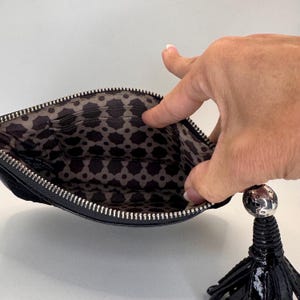 May include: Black leather clutch bag with a silver zip and a decorative tassel. The interior has a patterned fabric lining. The bag is open, showing the inside pockets.