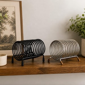 May include: Two metal letter holders, one black and one silver, sit on a wooden shelf. Each holder has a circular design with a base and small feet. A framed picture and a potted plant are in the background.