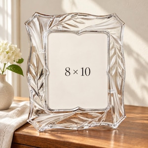May include: A clear glass picture frame with a decorative leaf pattern etched into the border. The frame features a scalloped edge and holds an 8 x 10 inch insert. The design is transparent with subtle frosted details along the carved leaves.