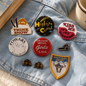 May include: A collection of eight assorted pins and patches featuring various designs, including text that reads Think Snow, Halley's Comet, Kiss Me It's My Birthday, Powars Because We Care, I Know What Girls Like, and Half Fast Skier, arranged on blue denim.