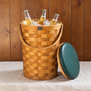 May include: A tall, cylindrical woven wood basket with a swing handle, containing three glass bottles filled with a golden liquid and ice. A separate round lid with a dark green padded top sits beside the basket on the surface.