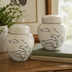 May include: Two off-white ceramic ginger jars with lids, each featuring a hand-painted black ink landscape design of a stylized tree and distant hills. The jars have a smooth, glossy finish and a rounded, bulbous shape with a short neck.