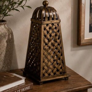 May include: A bronze-colored lantern with a woven design. The lantern has a domed top and a rectangular base. The woven pattern creates a lattice effect. The lantern is sitting on a wooden surface next to a book that says "CALL IT HOME".