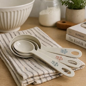 May include: A set of four off-white measuring spoons with printed measurements in red and blue. The spoons are arranged on a striped linen towel. A large white bowl and other kitchen items are in the background.