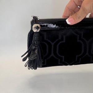 May include: Black clutch bag with a geometric pattern and a shiny, patent leather tassel. The bag has a silver zip and a decorative silver charm. The bag is made of a soft, velvet-like material.