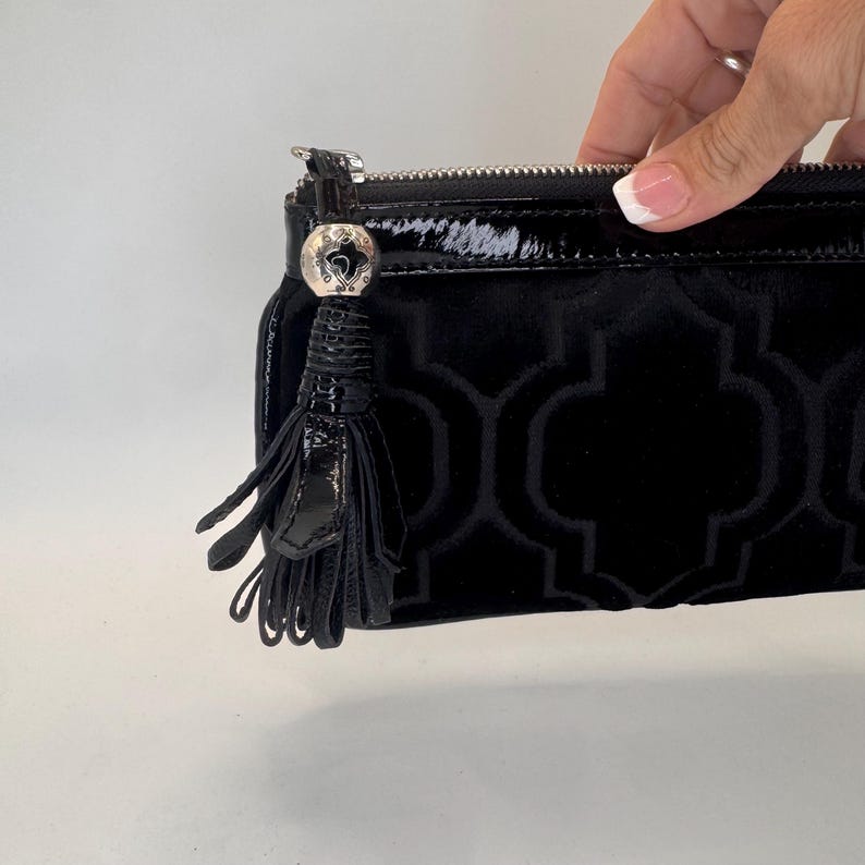 May include: Black clutch bag with a geometric pattern and a shiny, patent leather tassel. The bag has a silver zip and a decorative silver charm. The bag is made of a soft, velvet-like material.