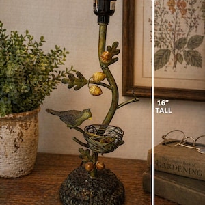 Puede incluir: Lámpara de mesa decorativa con un diseño inspirado en la naturaleza. La lámpara presenta un pájaro posado en una rama con un nido, bellotas y una base texturizada. Mide 40.6 cm de alto y tiene una base de 12.7 cm. La pantalla es beige.