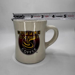 May include: Cream-colored ceramic coffee mug with the Waffle House Coffee logo in red, yellow, and black. The mug has a curved handle and is approximately 3 inches tall.