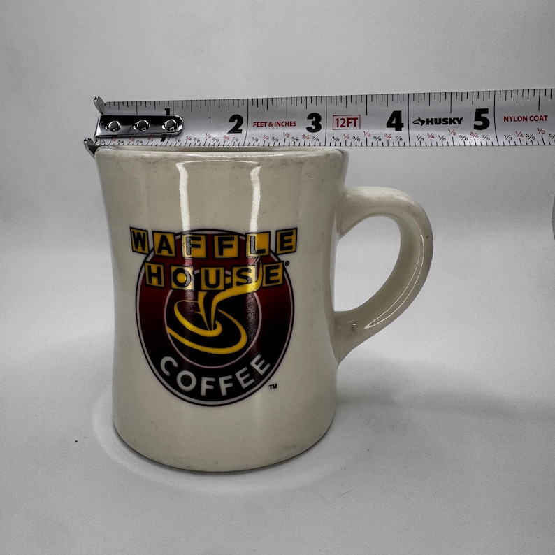 May include: Cream-colored ceramic coffee mug with the Waffle House Coffee logo in red, yellow, and black. The mug has a curved handle and is approximately 3 inches tall.