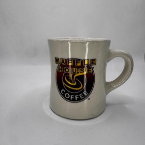 May include: Cream-colored ceramic coffee mug with the Waffle House logo. The logo features the words "WAFFLE HOUSE" in yellow and black, and "COFFEE" in white, with a swirl design in the center. The mug has a curved handle.