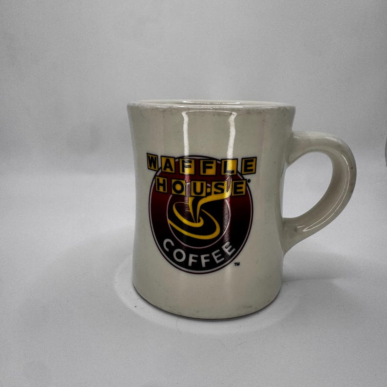 May include: Cream-colored ceramic coffee mug with the Waffle House logo. The logo features the words "WAFFLE HOUSE" in yellow and black, and "COFFEE" in white, with a swirl design in the center. The mug has a curved handle.