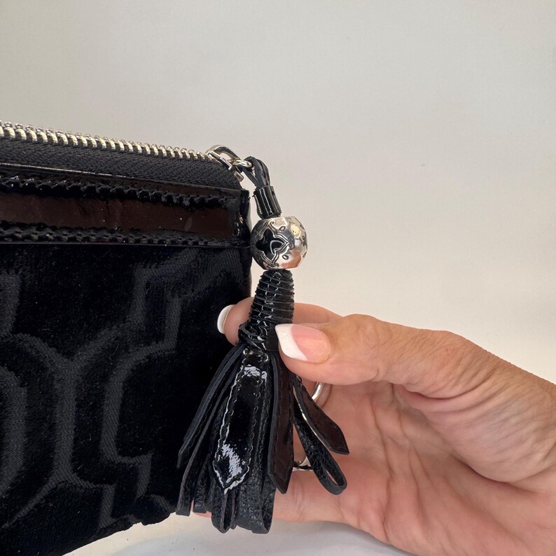 May include: Black velvet clutch with a geometric pattern. The clutch has a shiny black patent leather trim and a silver zipper. A black tassel with a silver bead hangs from the zipper.