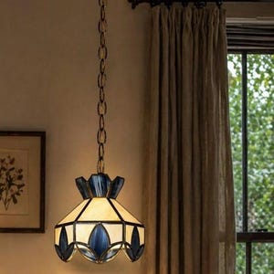 Op de afbeelding: Een hanglamp met een gebrandschilderde lampenkap. De kap heeft een geometrisch ontwerp met crème, blauwe en heldere glazen panelen. Hij hangt aan een messing ketting, wat een vleugje vintage aan de kamer toevoegt.