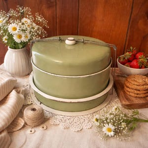 Könnte beinhalten: Ein zweistufiger, salbeigrüner Kuchenbehälter mit weißer Umrandung und einem Metallgriff. Der Behälter steht auf einer weißen Spitzendecke, daneben eine Vase mit Gänseblümchen und eine Schüssel mit Erdbeeren. Ein Stapel Kekse liegt auf einem Holzbrett.