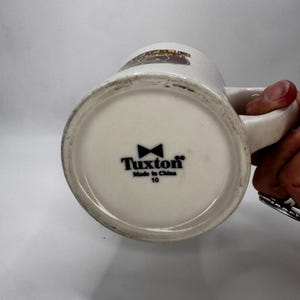 May include: A white ceramic mug with a handle, featuring the text "Tuxton" and a bow tie logo on the bottom. The mug has a slightly off-white color and a design on the top.
