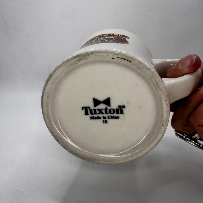 May include: A white ceramic mug with a handle, featuring the text "Tuxton" and a bow tie logo on the bottom. The mug has a slightly off-white color and a design on the top.