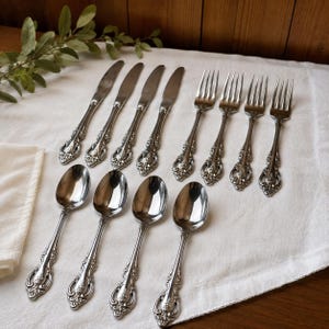 May include: A set of twelve stainless steel flatware pieces including four knives, four forks, and four spoons. Each piece features a polished silver finish and an ornate, embossed floral scrollwork pattern along the handle base and neck.