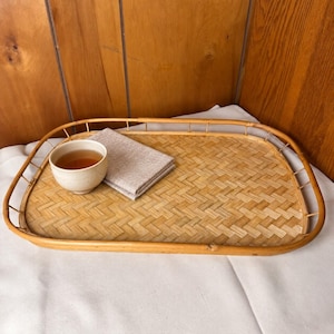 May include: A rectangular woven rattan tray with a raised border and a light brown finish. A small, off-white ceramic cup filled with a brown liquid and a folded, light beige linen napkin are placed on the surface of the tray.