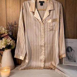 May include: A long-sleeved button-down pyjama shirt in a champagne-coloured satin fabric with thin vertical dark stripes. The shirt features a notched collar, a single chest pocket, and cuffs on the sleeves. A black label at the neck reads HALSTON.