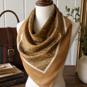 May include: A square silk scarf in a golden brown hue featuring a delicate green and white floral vine pattern. The scarf has a solid border with a white stripe and the word GUCCI printed in white at the bottom center, draped on a mannequin.