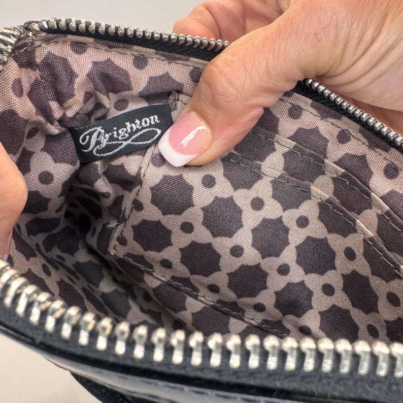 May include: Black leather wallet with a silver zip, open to show a patterned interior. The interior has a repeating geometric design in grey shades and a label that says "Brighton".