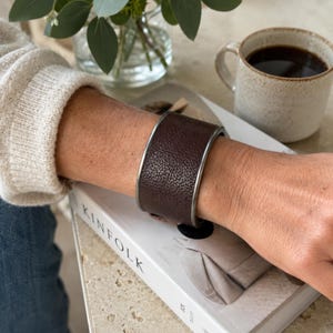 Puede incluir: Una pulsera de brazalete ancho con una banda de cuero texturizado de color marrón oscuro enmarcada por bordes de metal liso en tono plateado. La pulsera tiene una estructura cilíndrica sólida que envuelve la muñeca, mostrando un acabado de grano granulado distintivo en la superficie del cuero y un borde metálico limpio en los lados superior e inferior.
