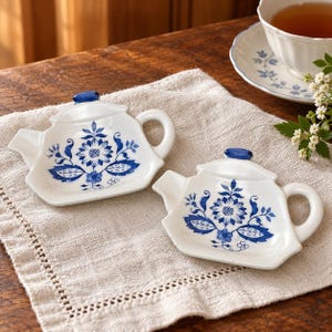 May include: Two white ceramic tea bag rests shaped like miniature teapots, each featuring a blue floral pattern and a blue handle on the lid. The small dishes have a spout, a handle, and a flat base for holding used tea bags.