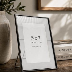 Pode incluir: Uma moldura de fotos preta de 5x7 (12,7 x 17,8 cm) com um passe-partout branco, exibida em uma superfície de madeira. A moldura é suportada por um suporte de metal. O texto na moldura diz "5 x 7 PHOTO FRAME 5.5 x 7.5 FRAME".