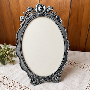 May include: An oval-shaped picture frame with a metallic silver finish. The frame features a textured border and is decorated with sculpted rose and leaf motifs at the top and bottom. The center contains a blank, off-white rectangular insert.