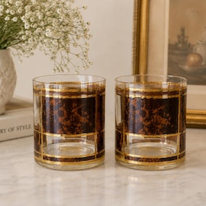 May include: Two clear, vintage-style drinking glasses with a brown and gold geometric design. The glasses have a rectangular pattern with a textured brown center. The glasses are on a marble surface.