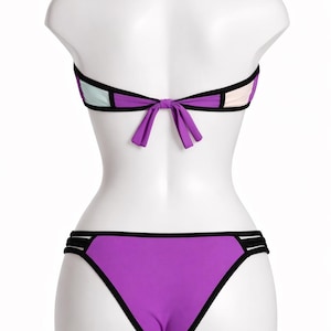 May include: A purple, black, and white bikini set on a mannequin. The bandeau top features a bow at the back, and the bottoms are a classic cut. The bikini has black trim and colour-blocked panels. A summer swimwear item.