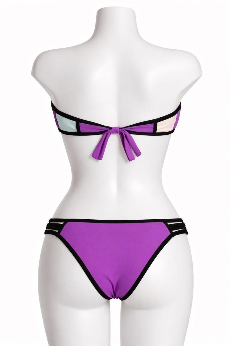 May include: A purple, black, and white bikini set on a mannequin. The bandeau top features a bow at the back, and the bottoms are a classic cut. The bikini has black trim and colour-blocked panels. A summer swimwear item.