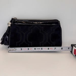 May include: Black clutch bag with a velvet geometric pattern and a glossy top. The clutch has a zip closure and a tassel. The clutch is approximately 20 cm long.