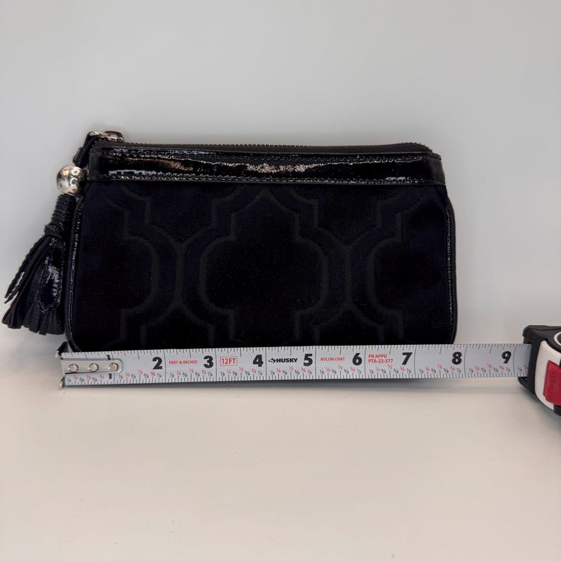 May include: Black clutch bag with a velvet geometric pattern and a glossy top. The clutch has a zip closure and a tassel. The clutch is approximately 20 cm long.