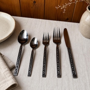 Puede incluir: Un juego de cinco cubiertos de acero inoxidable que incluye una cuchara sopera, una cucharilla, un tenedor de mesa, un tenedor de postre y un cuchillo. Cada pieza tiene un acabado metálico oscuro y un patrón geométrico en relieve en el mango.